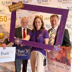 Drei Personen posieren in einem glitzernden Rahmen vor einem Banner, das das 50-j&auml;hrige Jubil&auml;um des TSC Br&uuml;hl feiert.