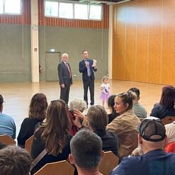 Zwei M&auml;nner stehen auf einer B&uuml;hne, w&auml;hrend ein kleines M&auml;dchen in einem lila Kleid neben ihnen an einer Veranstaltung teilnimmt.