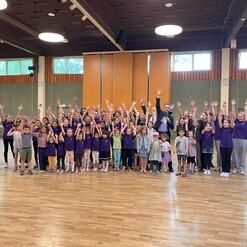 Gro&szlig;e Gruppe von Kindern und Erwachsenen in lila Shirts, die mit gehobenen H&auml;nden in einem Raum posieren.