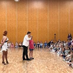 Tanzveranstaltung mit einem Paar auf der B&uuml;hne und einer Gruppe von Kindern und Erwachsenen, die im Publikum zuschauen.