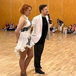 Eine tanzende Frau in wei&szlig;em Kleid und ein Mann im Anzug sprechen vor einer Zuschauergruppe in einem Holzsaal.