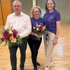 Drei Personen, die Blumenstr&auml;u&szlig;e halten, stehen in einer Halle mit Holzfu&szlig;boden.