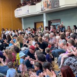 Vielz&auml;hlige Menschen applaudieren in einem Innenraum, versammelt auf Sitzen, mit Kindern und Erwachsenen in unterschiedlichen Altersgruppen.