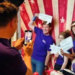 Personen in lila Shirts posieren mit Sprechblasen vor einem gestreiften Hintergrund, w&auml;hrend einer ein Foto aufnimmt.