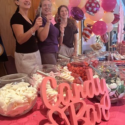Drei Frauen l&auml;cheln, halten L&ouml;ffel in der Hand, vor einer bunten Candy-Bar mit S&uuml;&szlig;igkeiten und Luftballons.