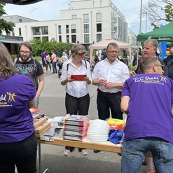 Menschen stehen an einem Stand des TSC Br&uuml;hl, umringt von Tischen mit B&uuml;chern und Spielwaren, bei einem Stra&szlig;enfest.