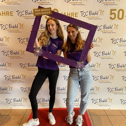 Zwei Frauen posieren mit einem lila Bilderrahmen vor einem Banner des TSC Br&uuml;hl, gekleidet in lila T-Shirts.