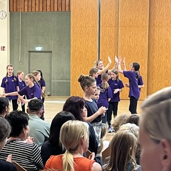Gruppe von M&auml;dchen in lila T-Shirts f&uuml;hrt eine Tanzauff&uuml;hrung in einem Saal vor Publikum auf.