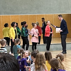 F&uuml;nf Frauen in bunten Outfits stehen auf einer B&uuml;hne, w&auml;hrend ein Mann sie interviewt. Zuschauer sitzen im Vordergrund.