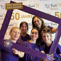 F&uuml;nf Frauen in lila T-Shirts posieren mit einem glitzernden Rahmen vor einem Banner zum 50-j&auml;hrigen Jubil&auml;um des TSC Br&uuml;hl.