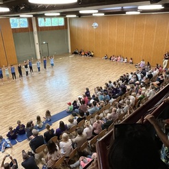 Tanzvorf&uuml;hrung mit mehreren T&auml;nzern auf einer Holzfl&auml;che, umgeben von einem Publikum auf Holzst&uuml;hlen.