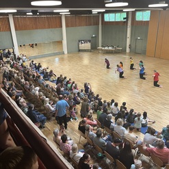 Tanzauff&uuml;hrung in einem gro&szlig;en Saal, umgeben von Zuschauern, mit T&auml;nzern in bunten Shirts auf dem Holzfu&szlig;boden.