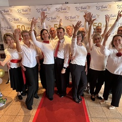 Gruppenselfie von T&auml;nzern in festlicher Kleidung, die vor einem Banner zum 50-j&auml;hrigen Jubil&auml;um posieren.