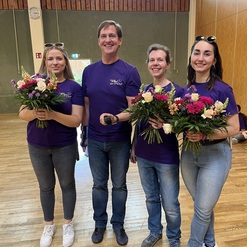 Vier Personen in lila T-Shirts halten Blumenstr&auml;u&szlig;e in einem Raum mit Holzboden. Im Hintergrund sind Fenster sichtbar.