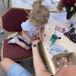 Ein kleines M&auml;dchen malt mit einem Pinsel ein Design auf den Arm eines Erwachsenen an einem Tisch.