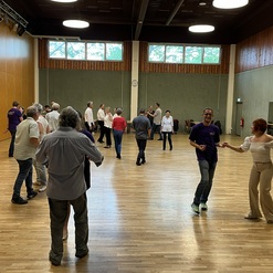 Tanzveranstaltung mit mehreren Paaren in einem gro&szlig;en Saal, umgeben von Holzvert&auml;felungen und gro&szlig;en Fenstern.