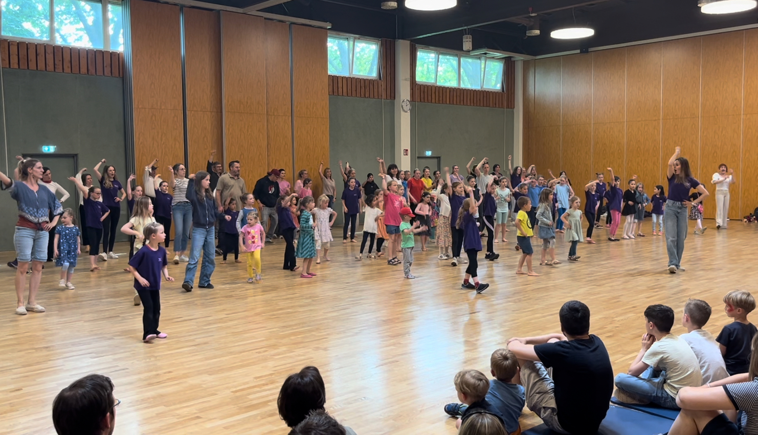 Versammlung von Kindern und Erwachsenen, die in einem hellen Raum gemeinsam tanzen und sich bewegen.