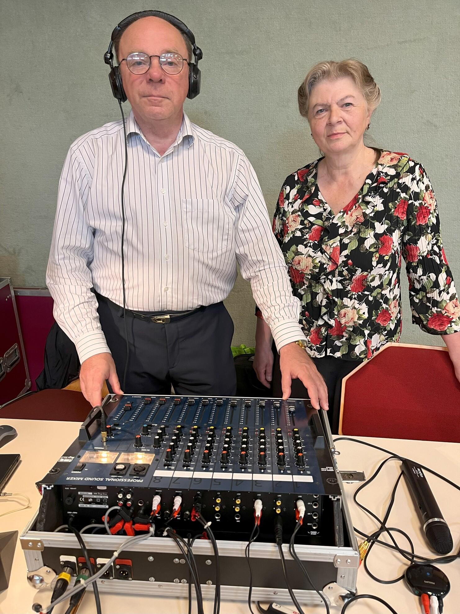 Zwei Personen stehen neben einem Audiomischpult, eins tr&auml;gt Kopfh&ouml;rer, beide blicken in die Kamera.