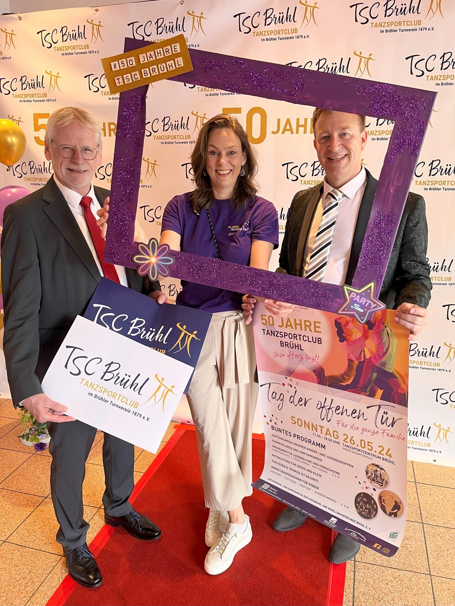 Drei Personen posieren vor einem Banner zur 50-Jahr-Feier des Tanzsportclubs Br&uuml;hl, halten Schilder und Plakate.