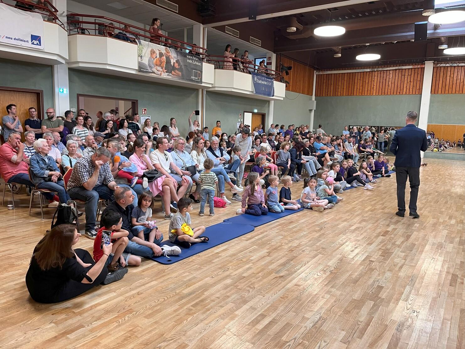 Gro&szlig;e Menschenmenge sitzt auf St&uuml;hlen und Matten in einem Saal, w&auml;hrend ein Redner auf der B&uuml;hne spricht.