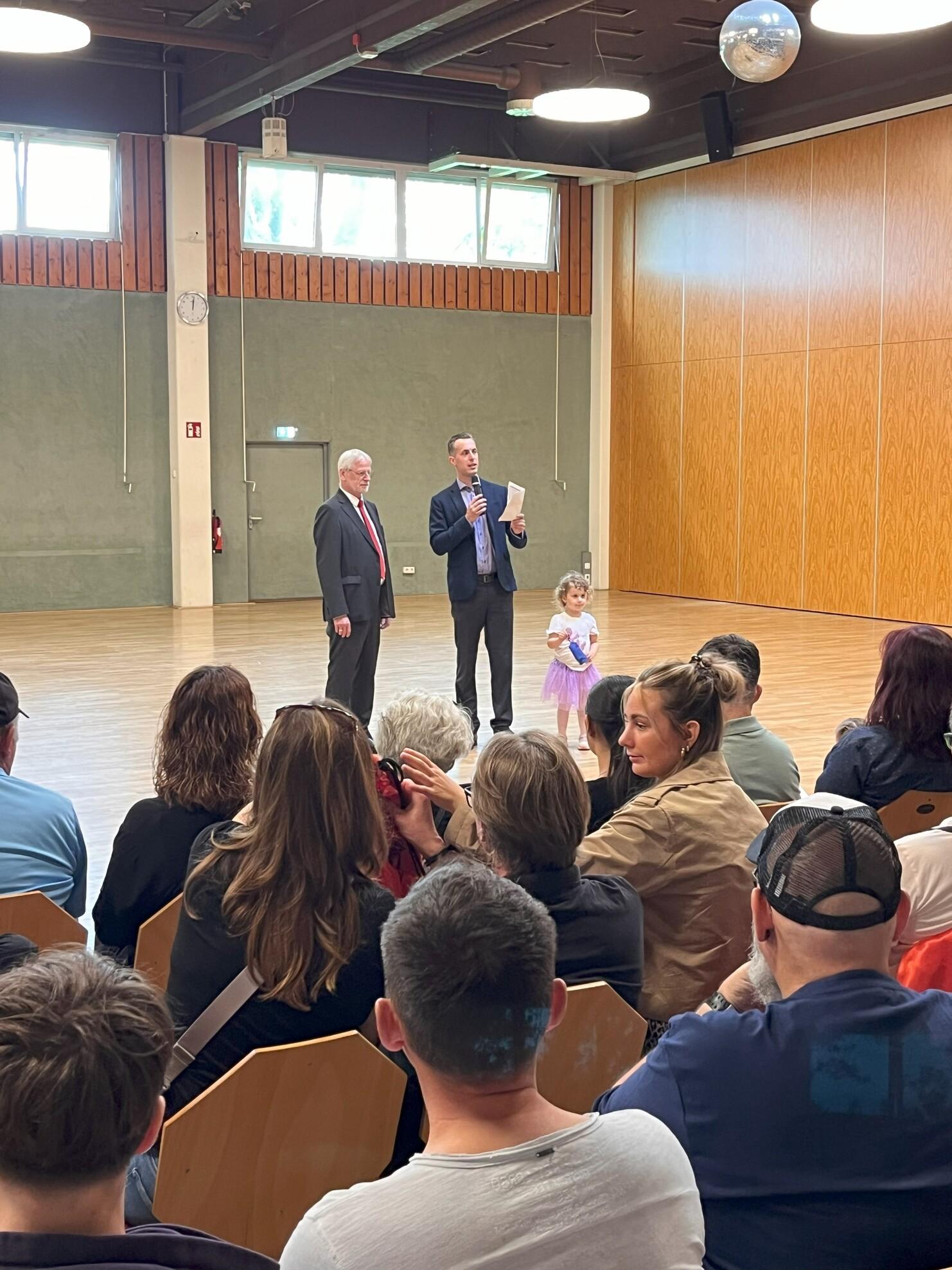 Zwei M&auml;nner stehen auf einer B&uuml;hne, w&auml;hrend ein kleines M&auml;dchen in einem lila Kleid neben ihnen an einer Veranstaltung teilnimmt.