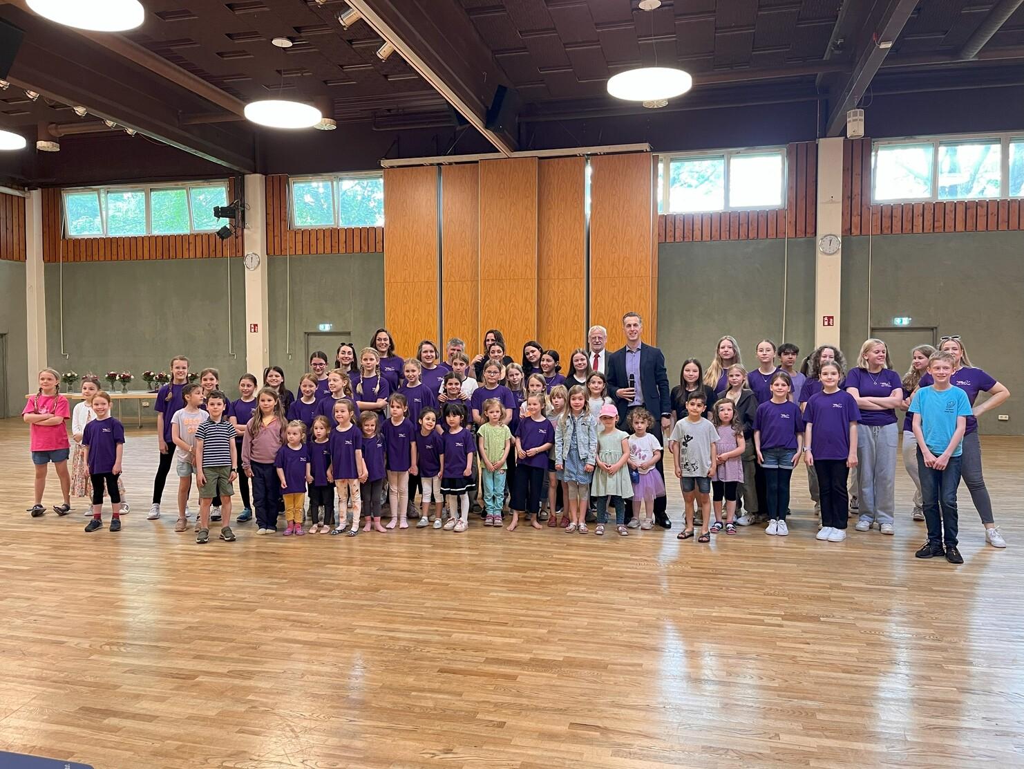 Gruppenfoto von Kindern und Erwachsenen in lila T-Shirts in einem gro&szlig;en Raum mit Holzfu&szlig;boden.