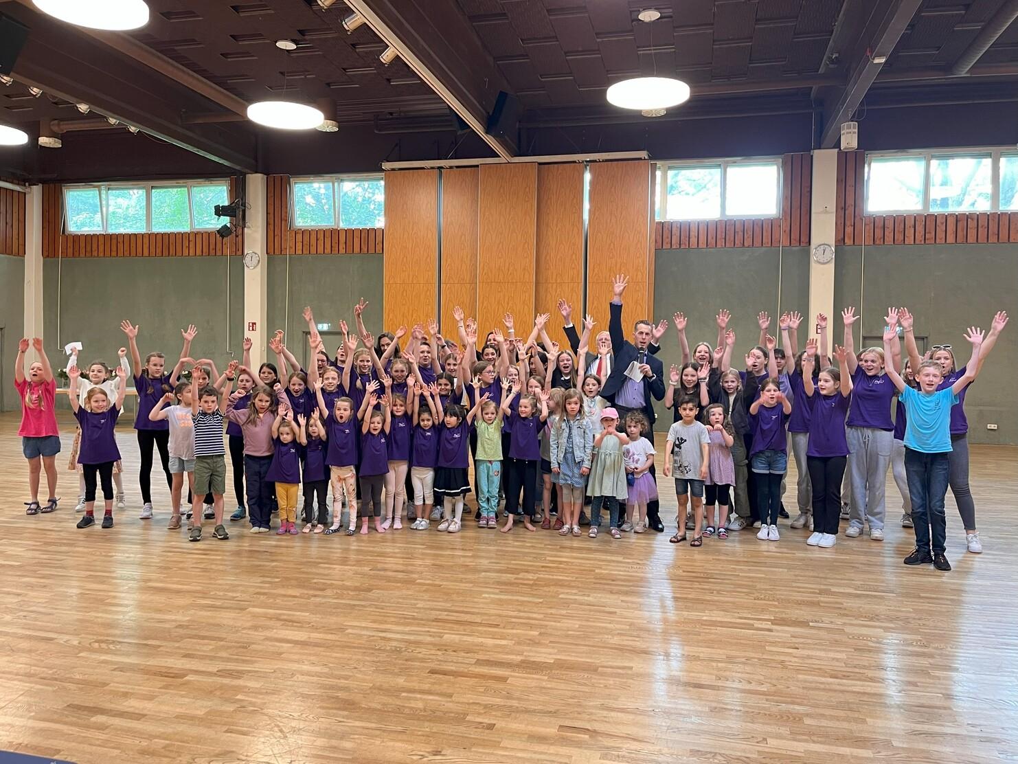 Gro&szlig;e Gruppe von Kindern und Erwachsenen in lila Shirts, die mit gehobenen H&auml;nden in einem Raum posieren.