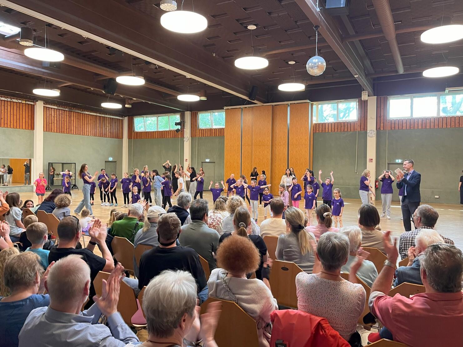 Tanzauff&uuml;hrung in einer Halle mit Kindern in lila Shirts, w&auml;hrend das Publikum applaudiert.