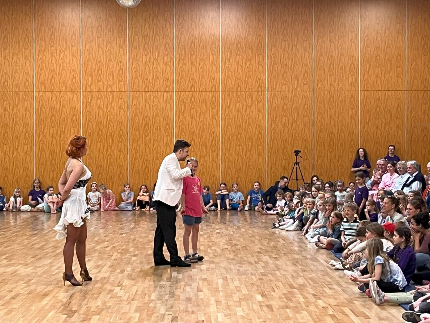 Tanzveranstaltung mit einem Paar auf der B&uuml;hne und einer Gruppe von Kindern und Erwachsenen, die im Publikum zuschauen.