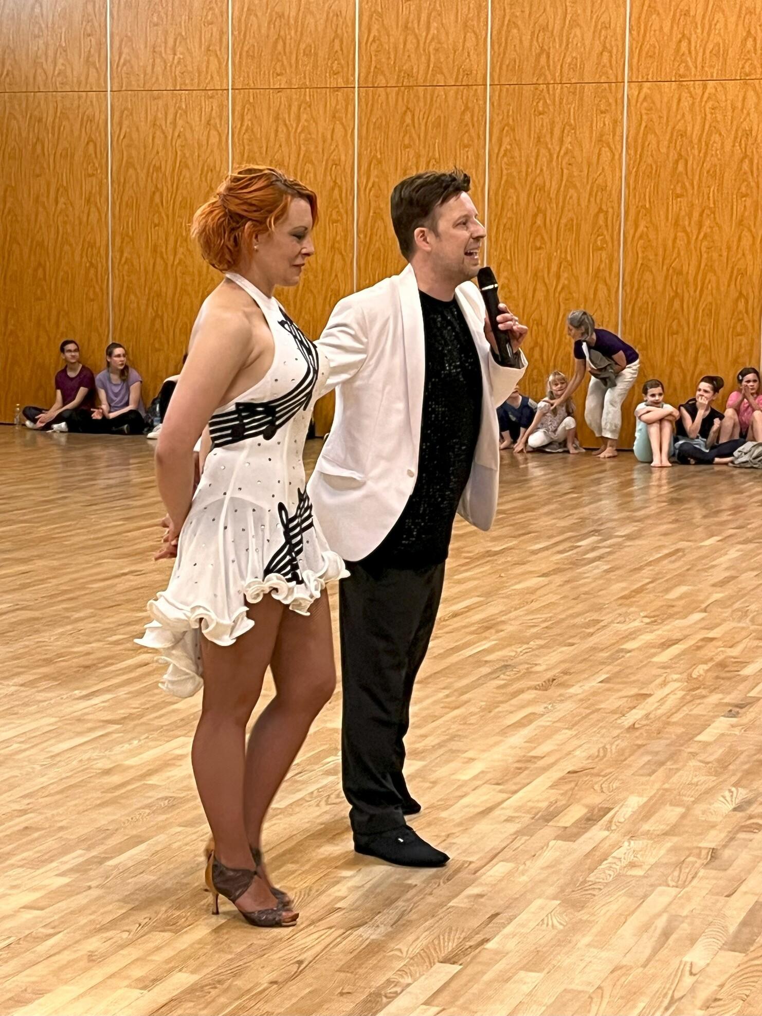 Eine tanzende Frau in wei&szlig;em Kleid und ein Mann im Anzug sprechen vor einer Zuschauergruppe in einem Holzsaal.