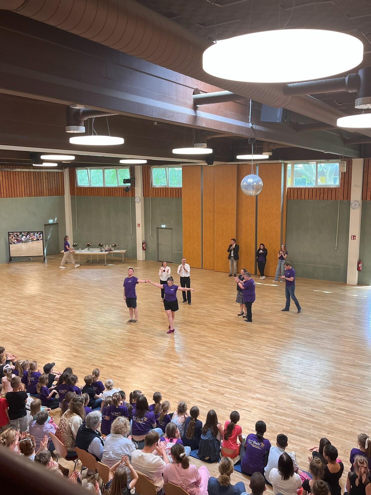 Tanzen auf einer B&uuml;hne: Teilnehmer in lila T-Shirts pr&auml;sentieren eine Choreografie vor einem applaudierenden Publikum.