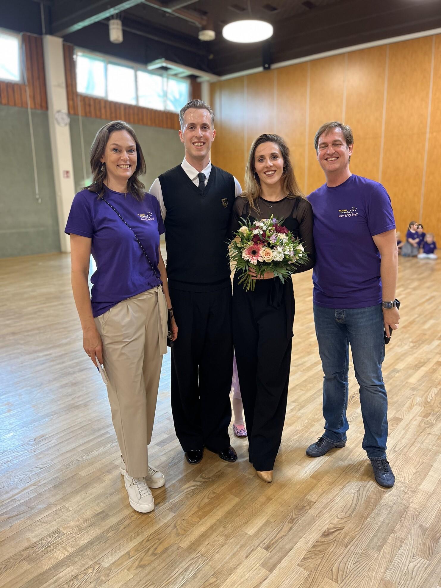 Vier Personen posieren in einem hellen Raum; zwei tragen lila T-Shirts, eine h&auml;lt einen Blumenstrau&szlig;.