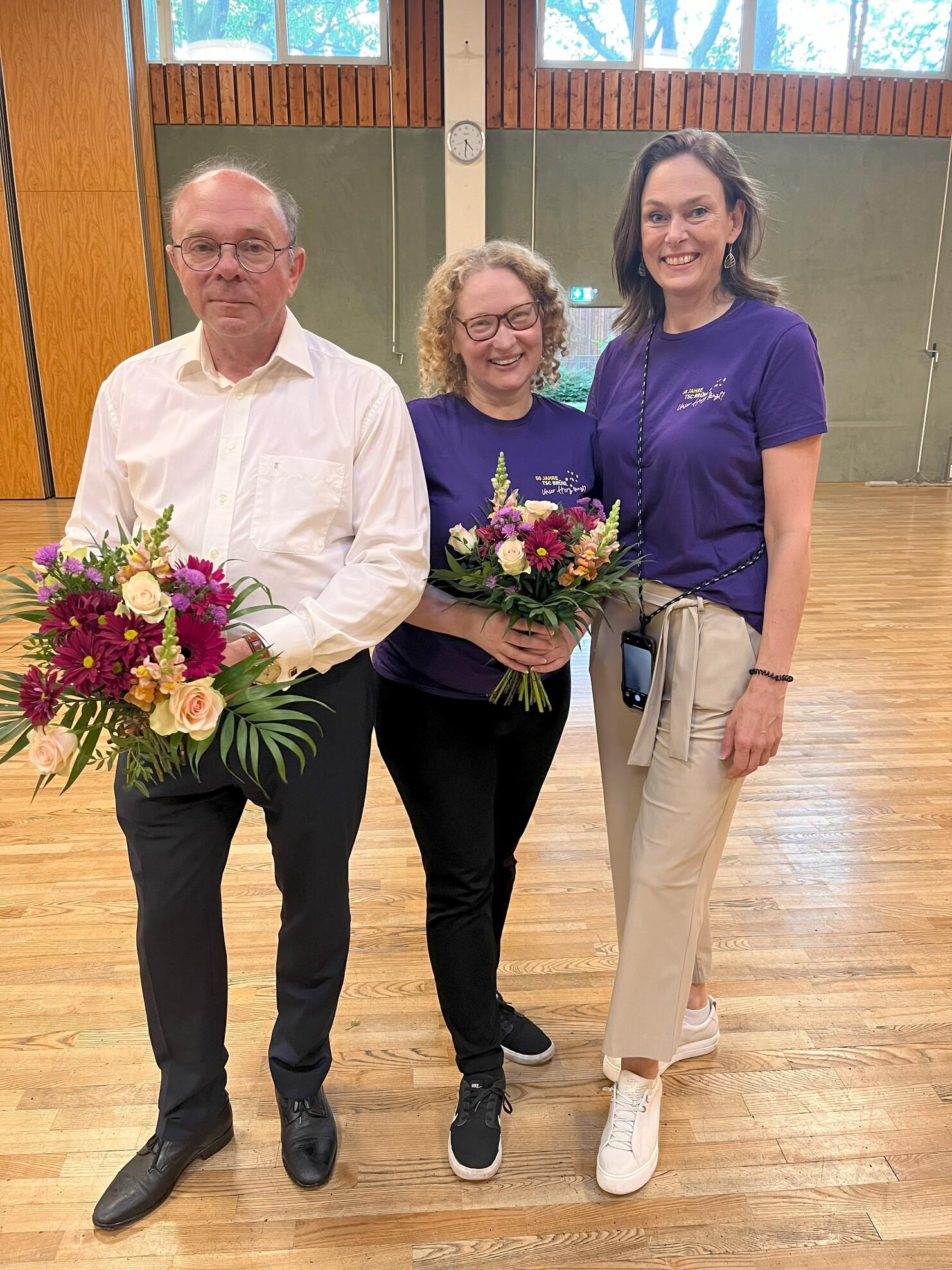 Drei Personen, die Blumenstr&auml;u&szlig;e halten, stehen in einer Halle mit Holzfu&szlig;boden.