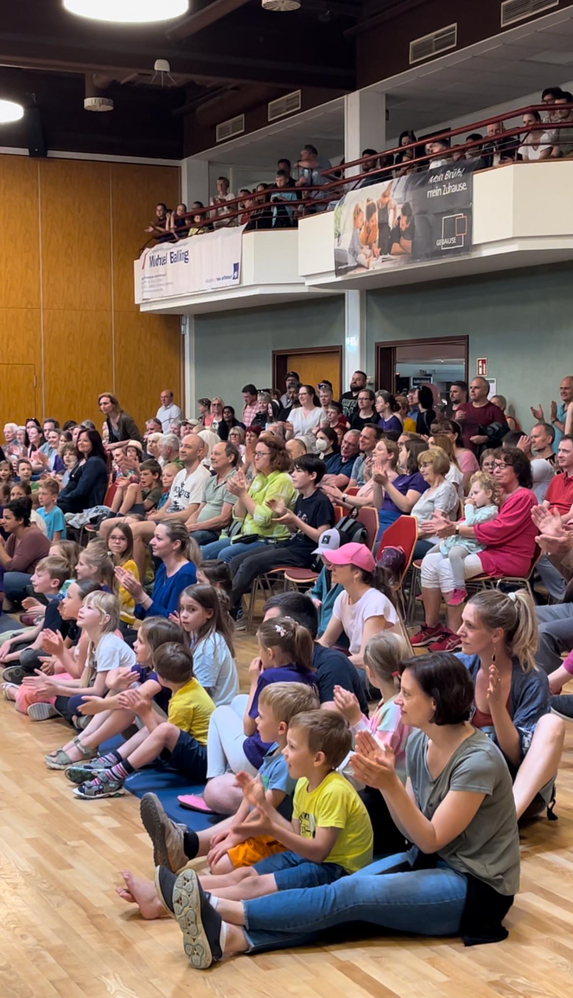 Eine gro&szlig;e Menge von Menschen, &uuml;berwiegend Kinder, sitzt auf dem Boden und applaudiert in einem Veranstaltungsraum.