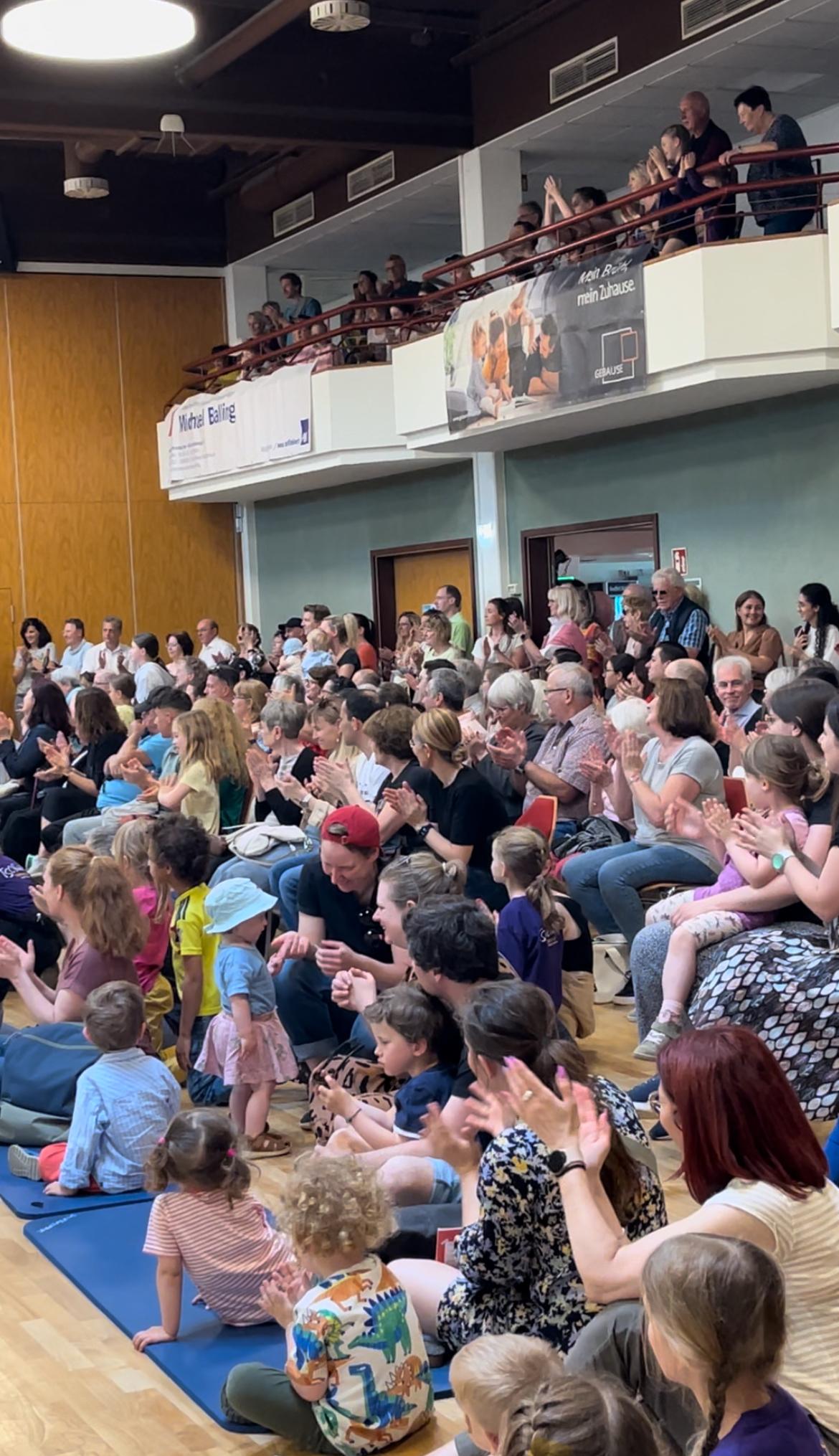 Vielz&auml;hlige Menschen applaudieren in einem Innenraum, versammelt auf Sitzen, mit Kindern und Erwachsenen in unterschiedlichen Altersgruppen.