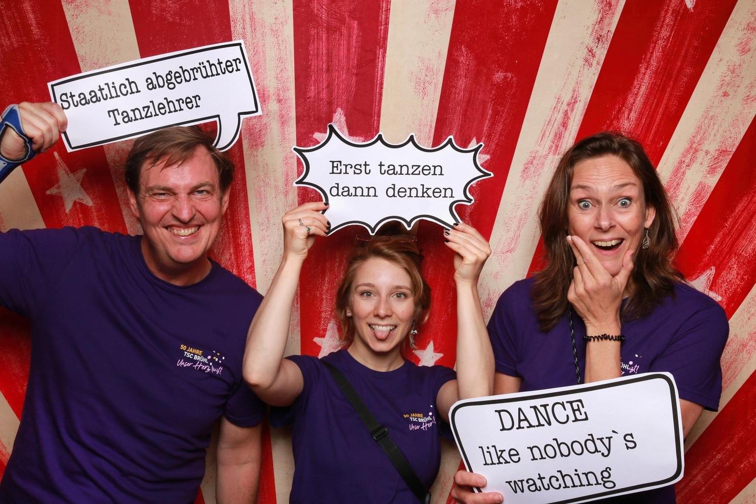 Drei Personen in lila Shirts halten Sprechblasen mit Spr&uuml;chen vor einem gestreiften Hintergrund.