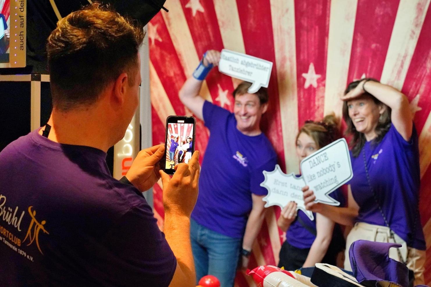 Personen in lila Shirts posieren mit Sprechblasen vor einem gestreiften Hintergrund, w&auml;hrend einer ein Foto aufnimmt.