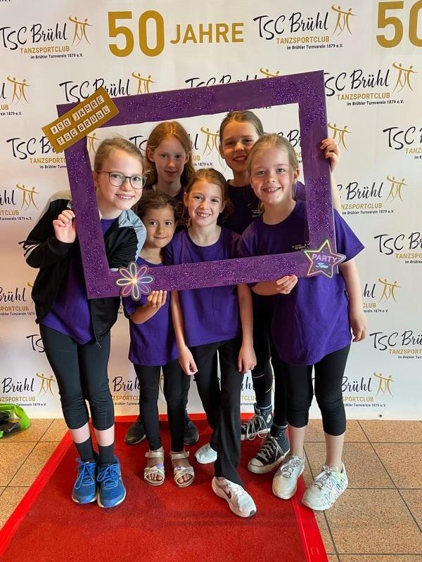 Sechs Kinder in lila T-Shirts posieren mit einem glitzernden Rahmen vor einem Banner zum 50-j&auml;hrigen Jubil&auml;um des TSC Br&uuml;hl.