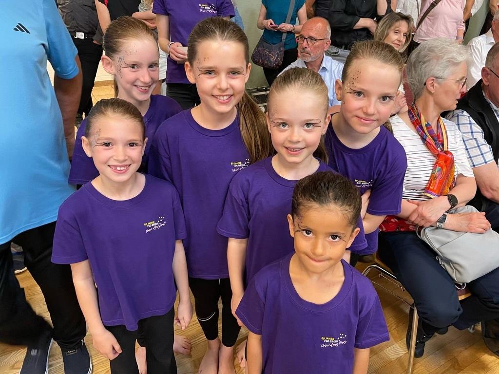 Sechs M&auml;dchen in lila T-Shirts mit Glitzerschminke posieren l&auml;chelnd in einer Menschenmenge.