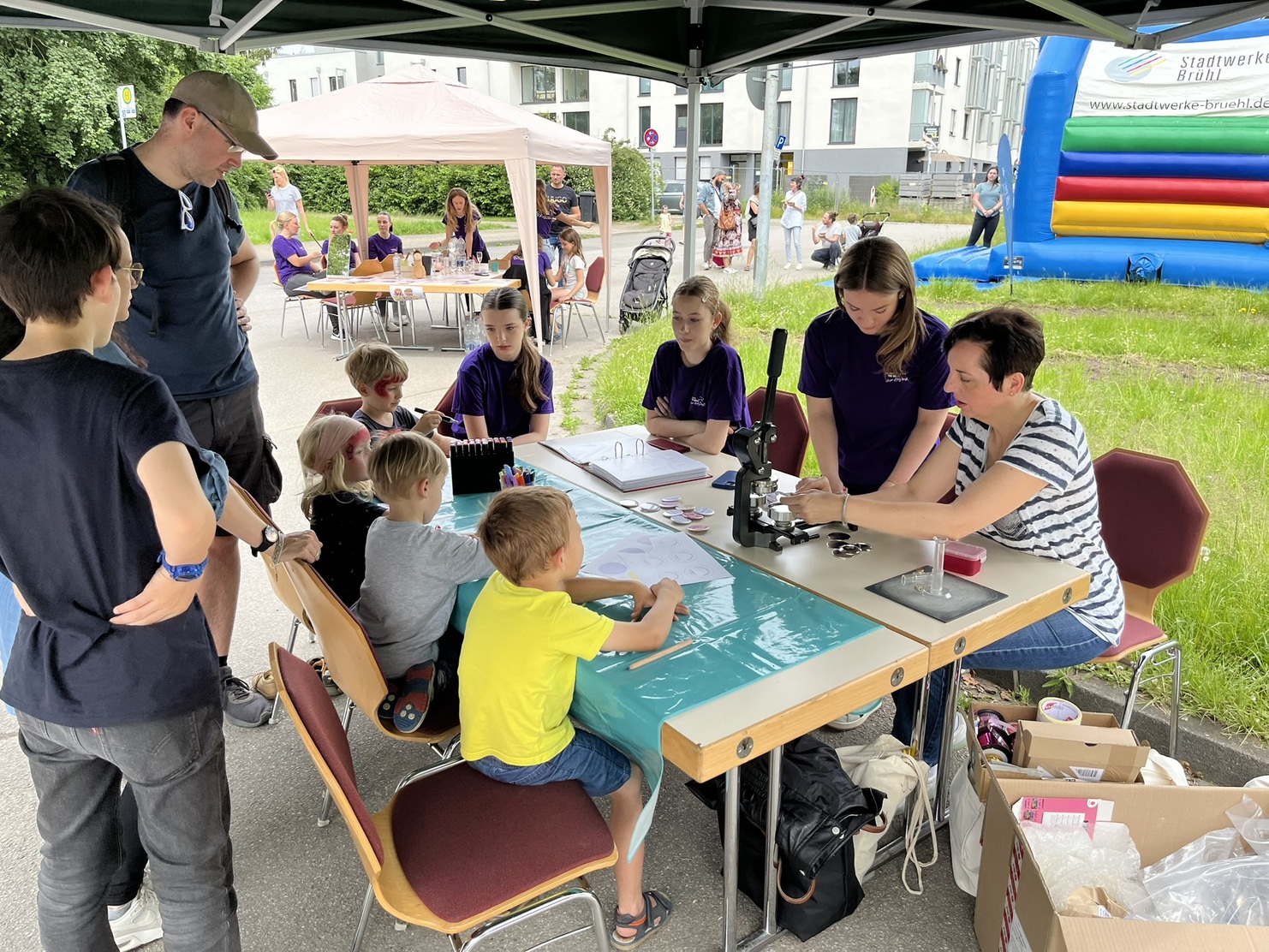 Kinder und Erwachsene am Basteltisch mit Mikroskop und Farben unter einem Zelt auf einer Au&szlig;eneventfl&auml;che.