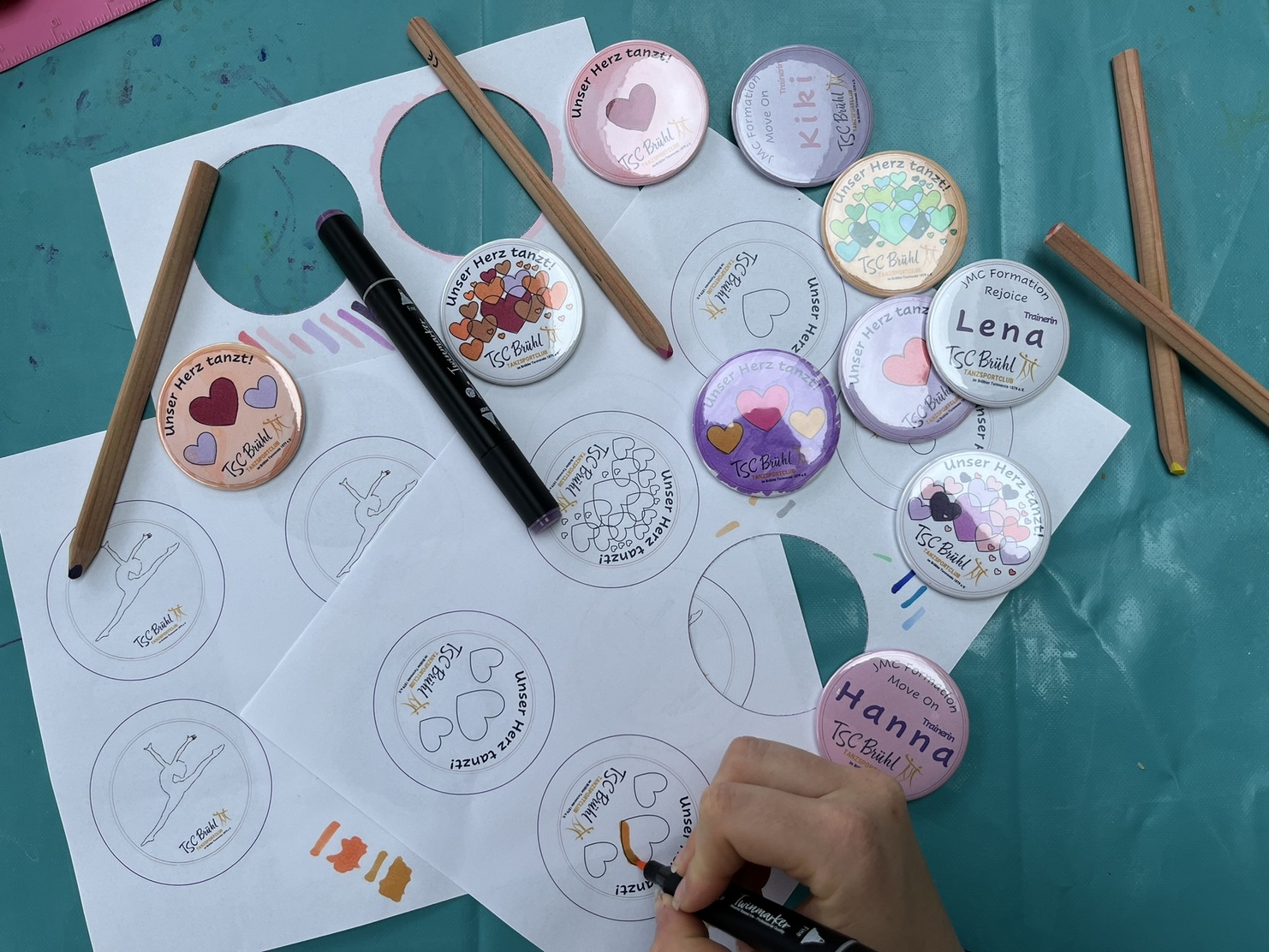 Bunt bedruckte Buttons und Zeichenskizzen liegen auf einem Tisch, w&auml;hrend jemand mit einem Marker zeichnet.