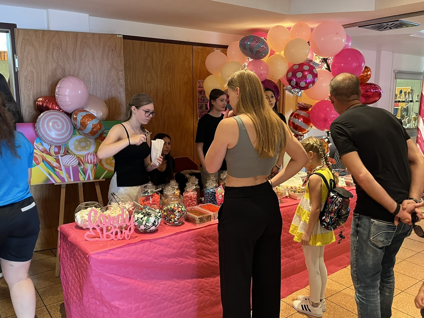 Bunte Candy-Bar mit S&uuml;&szlig;igkeiten und G&auml;sten, darunter ein Kind in gelbem Outfit und Luftballons im Hintergrund.