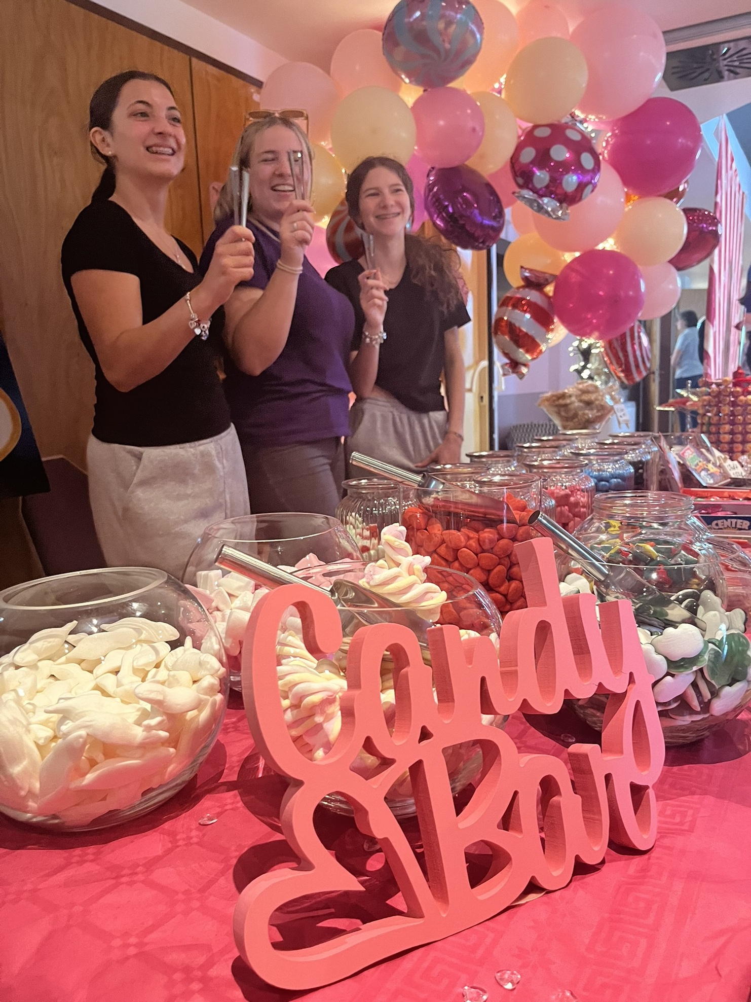 Drei Frauen l&auml;cheln, halten L&ouml;ffel in der Hand, vor einer bunten Candy-Bar mit S&uuml;&szlig;igkeiten und Luftballons.
