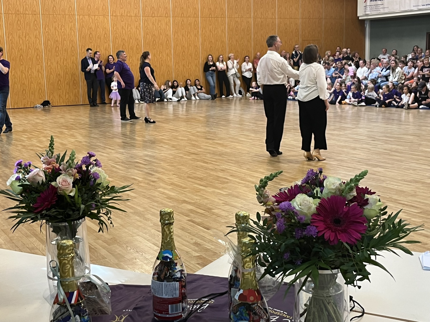 Tanzvorf&uuml;hrung in einem Festsaal mit Publikum, Fokus auf tanzendes Paar und Blumendekoration auf einem Tisch.