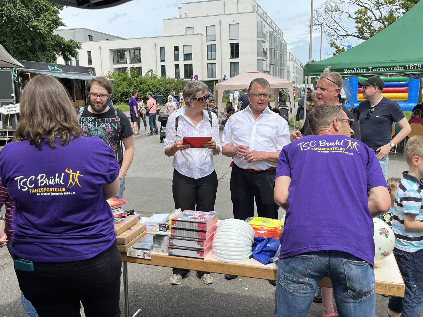 Menschen stehen an einem Stand des TSC Br&uuml;hl, umringt von Tischen mit B&uuml;chern und Spielwaren, bei einem Stra&szlig;enfest.