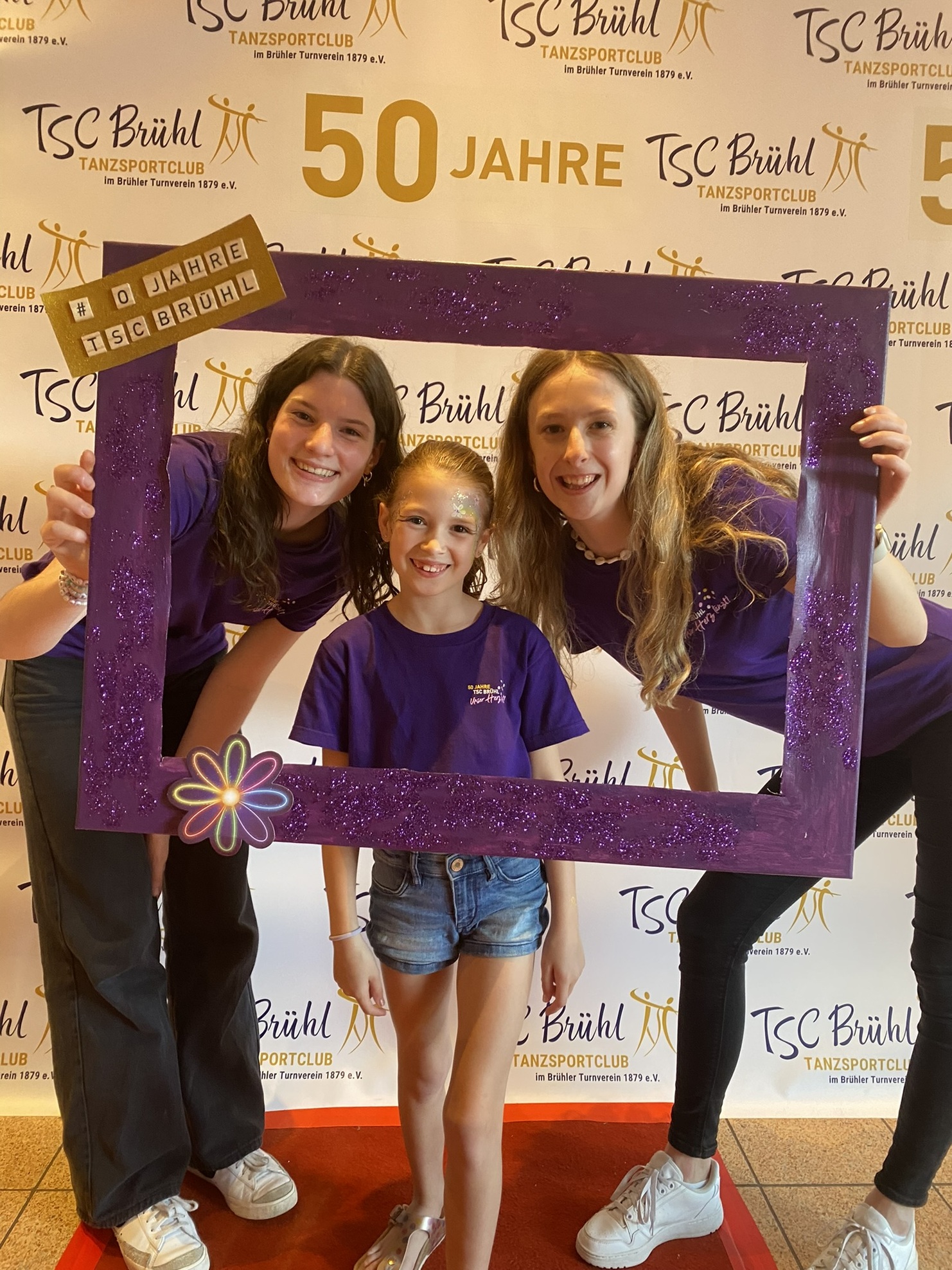 Drei Personen posieren in lila Shirts vor einer festlichen Hintergrundwand zum 50-j&auml;hrigen Jubil&auml;um des TSC Br&uuml;hl.