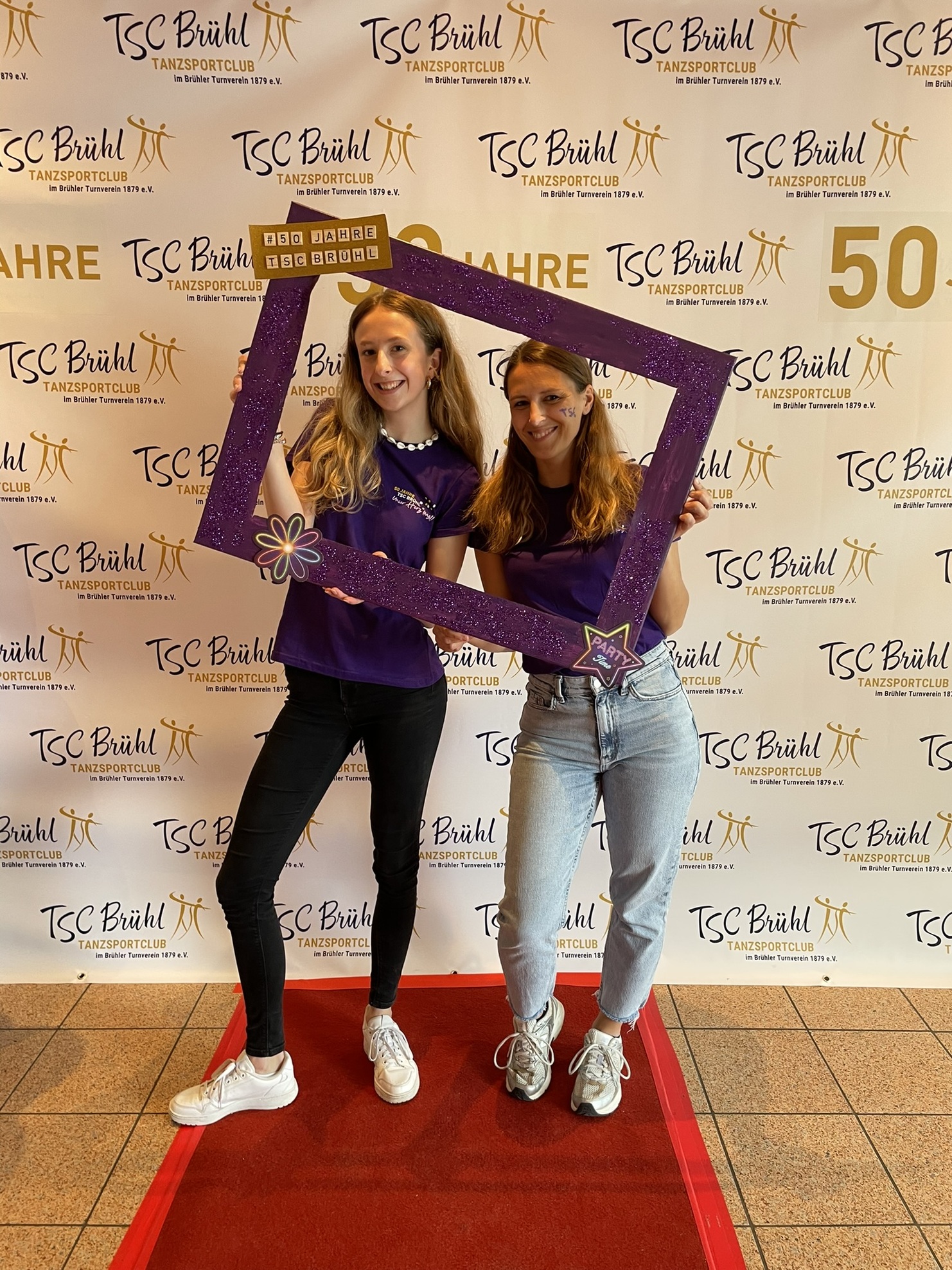 Zwei Frauen posieren mit einem lila Bilderrahmen vor einem Banner des TSC Br&uuml;hl, gekleidet in lila T-Shirts.