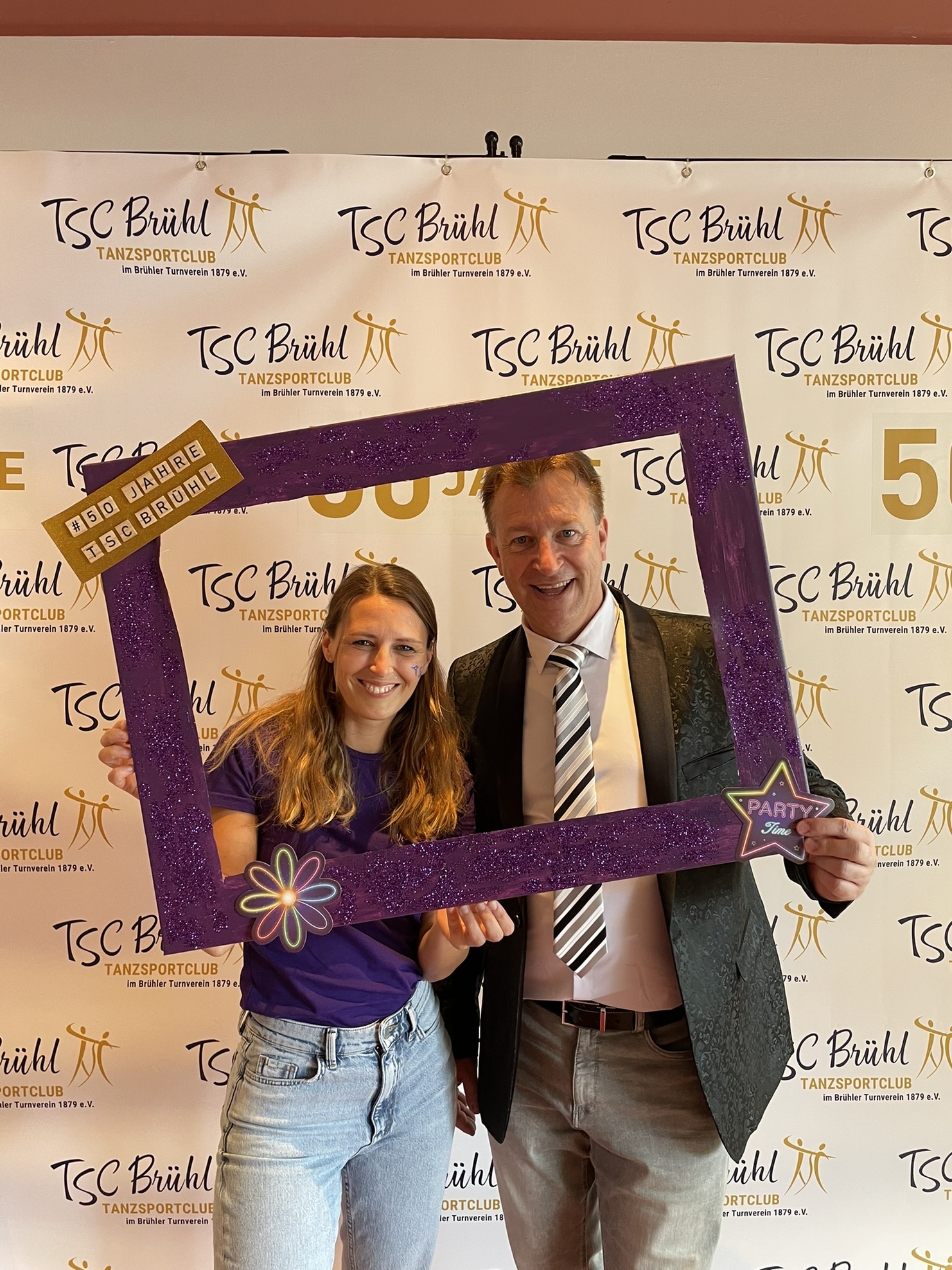 Junge Frau und Mann posieren l&auml;chelnd mit einem glitzernden Fotorahmen vor einem TSC Br&uuml;hl Jubil&auml;umsbanner.