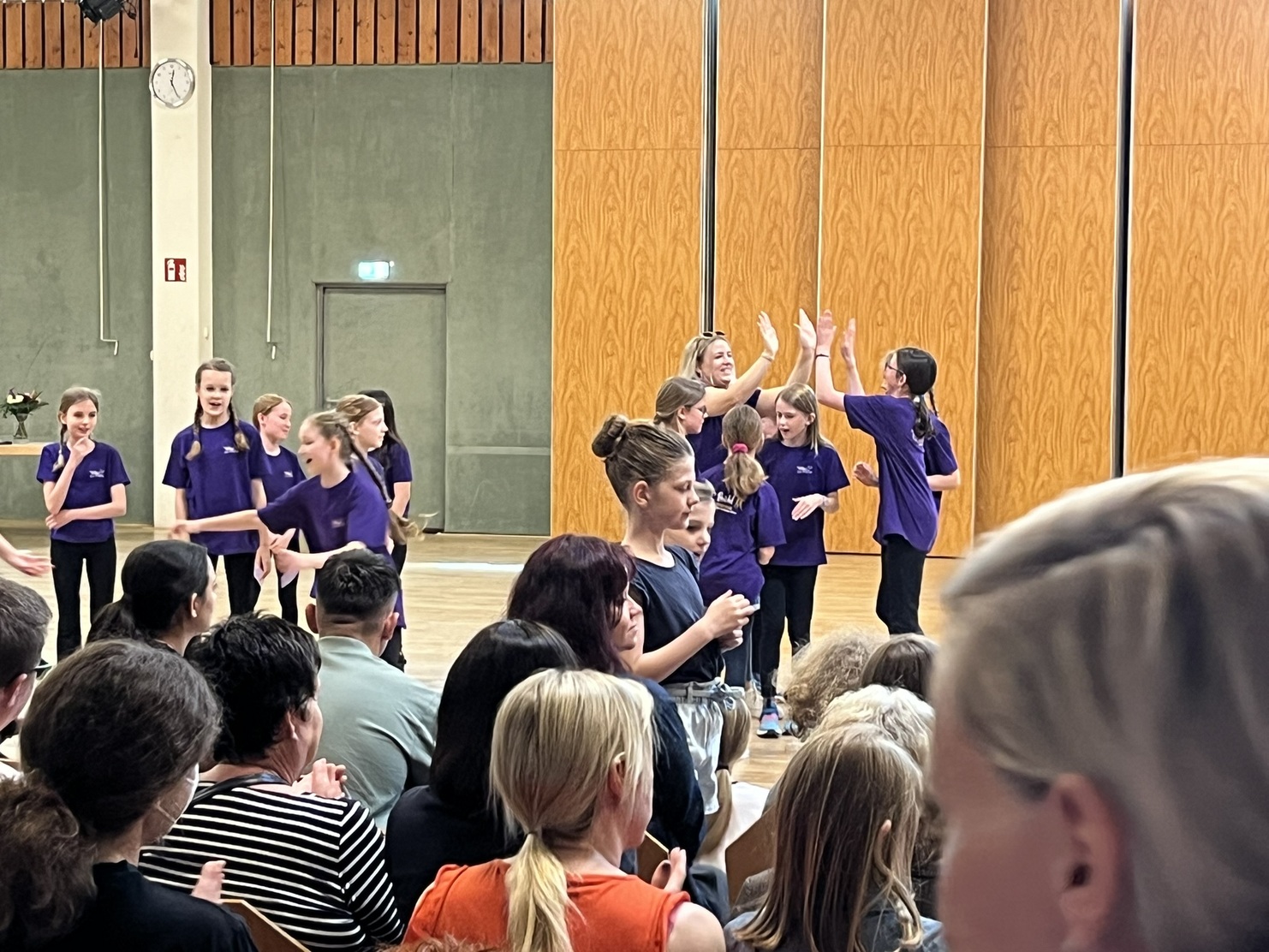 Gruppe von M&auml;dchen in lila T-Shirts f&uuml;hrt eine Tanzauff&uuml;hrung in einem Saal vor Publikum auf.
