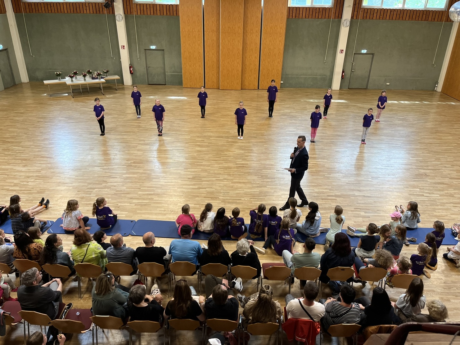 Eine Gruppe von T&auml;nzern in lila T-Shirts f&uuml;hrt eine Auff&uuml;hrung in einem gro&szlig;en Saal vor aufmerksamen Zuschauern auf.